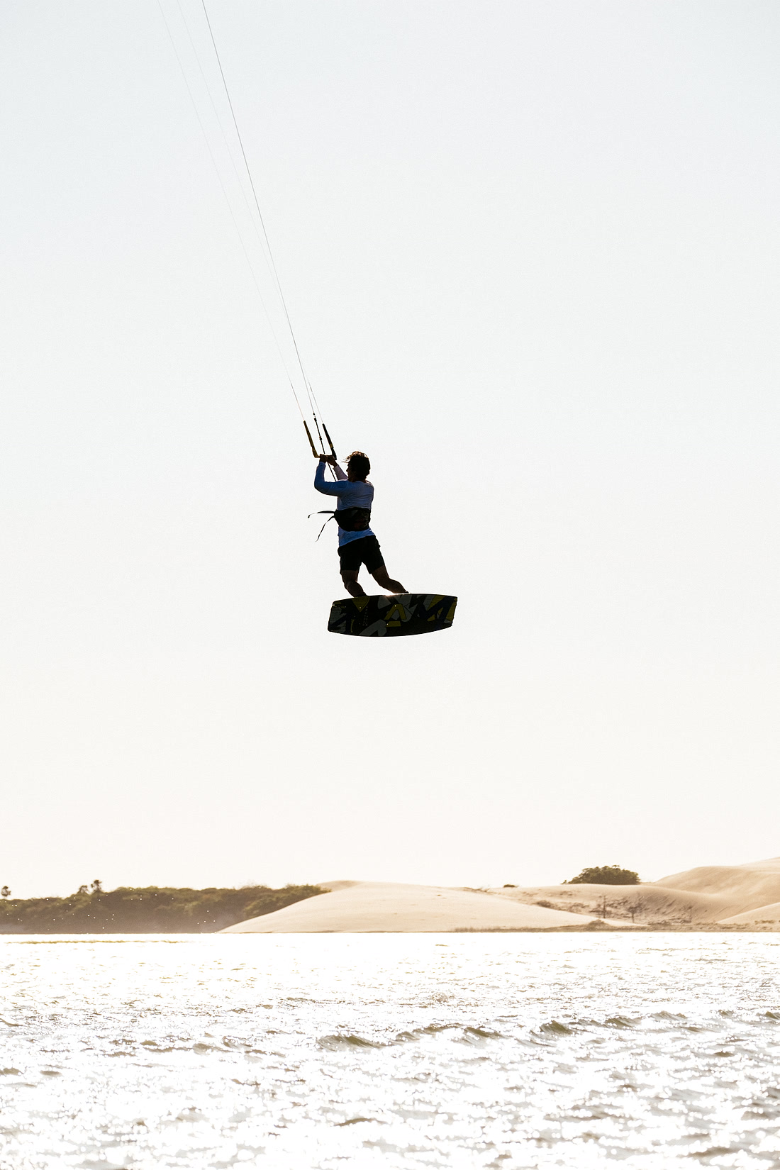 Kitesurfista em salto sobre as águas, com as dunas do litoral do Nordeste ao fundo